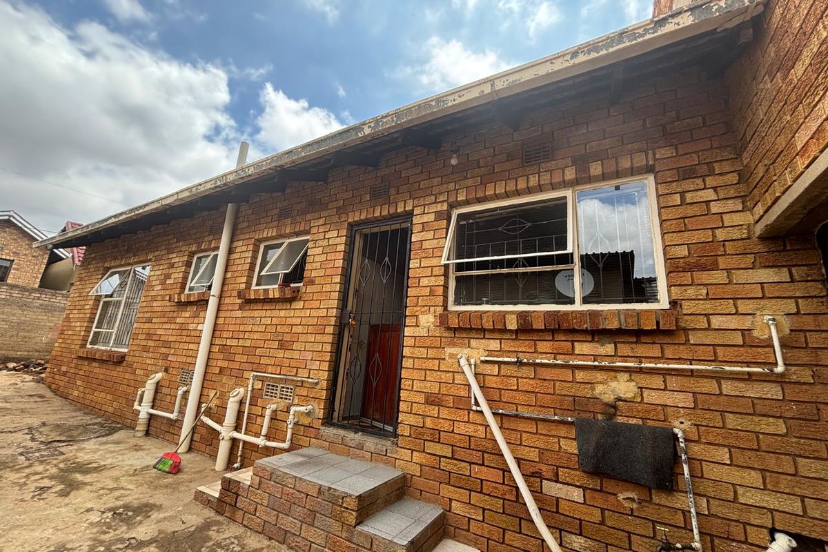 Rear exterior view of a face-brick house featuring exposed plumbing, security gates, and tiled entrance steps.