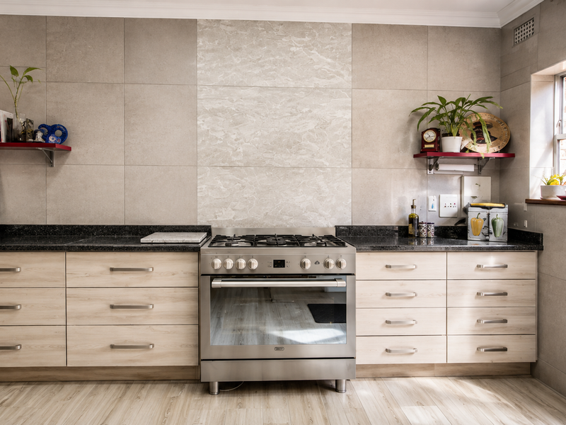 Modern kitchen featuring light wood-finish cabinetry, granite countertops, a stainless steel gas stove, and large format wall tiles with a decorative marble-look feature panel.