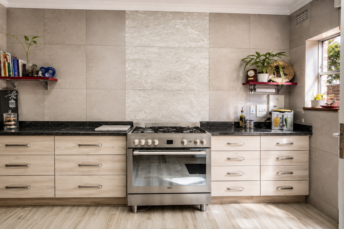 Modern kitchen featuring light wood-finish cabinetry, granite countertops, a stainless steel gas stove, and large format wall tiles with a decorative marble-look feature panel.