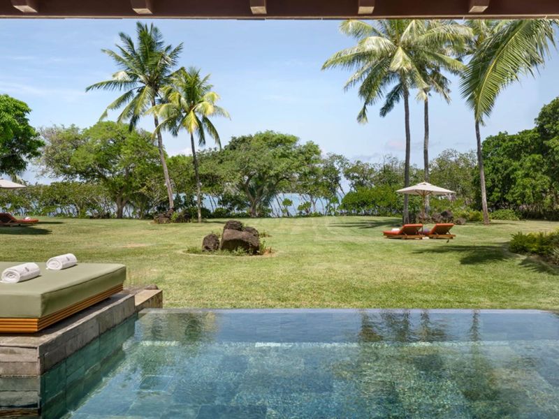 A serene outdoor pool area featuring a stone-tiled swimming pool overlooking a lush, expansive garden with mature palm trees and coastal views.