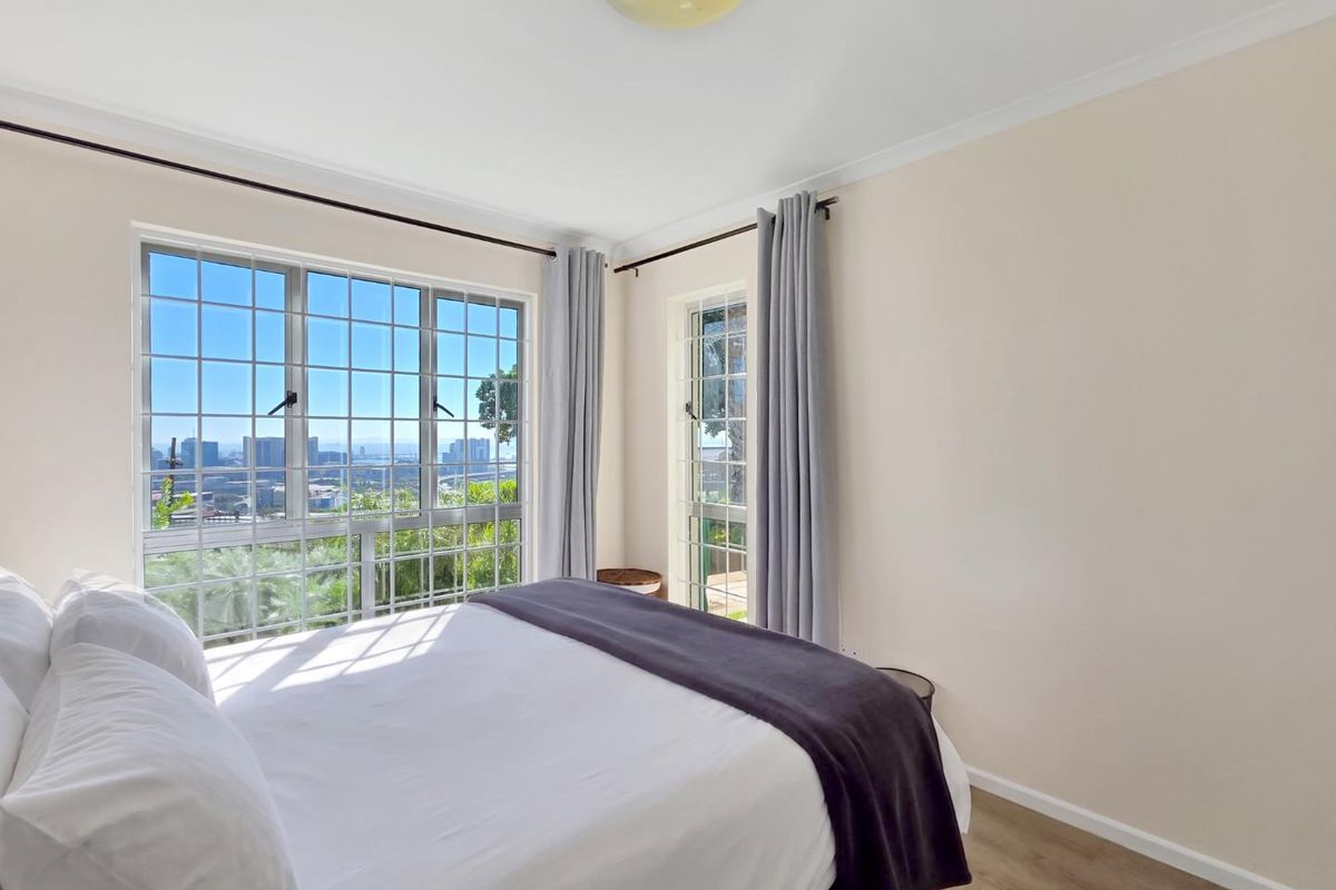 A bright bedroom featuring large grid-pattern windows with expansive city views, neutral walls, and modern laminate flooring.