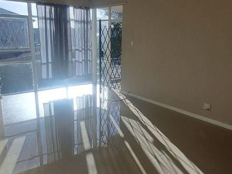 A bright, empty living room featuring tiled flooring, a ceiling light fixture, and a sliding glass door leading to an outdoor area with security gates.