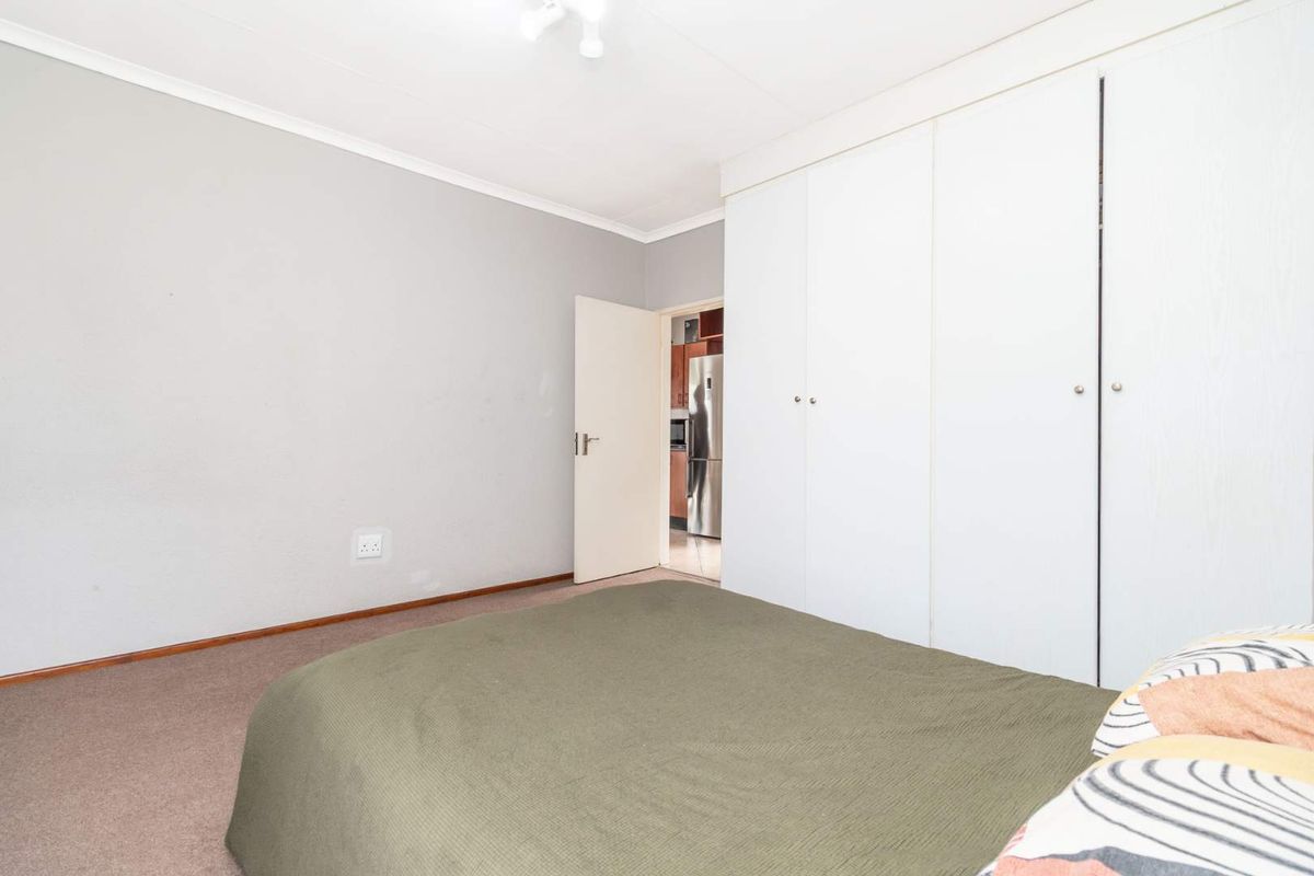 A bedroom featuring built-in cupboards with white doors, neutral wall finishes, and carpeted flooring, with a view into an adjacent kitchen area.