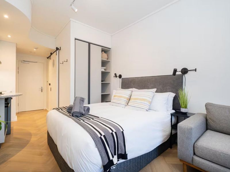 A modern open-plan studio apartment featuring herringbone timber flooring, a compact kitchen with stone countertops, and a built-in wardrobe.