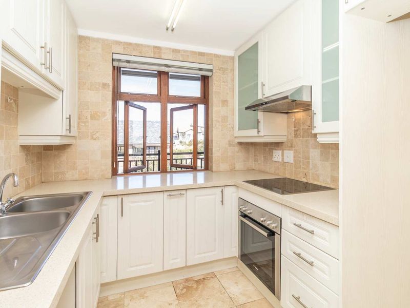 A clean, functional kitchen featuring white cabinetry, stone countertops, a double basin sink, and integrated appliances with natural light from a timber-framed window.