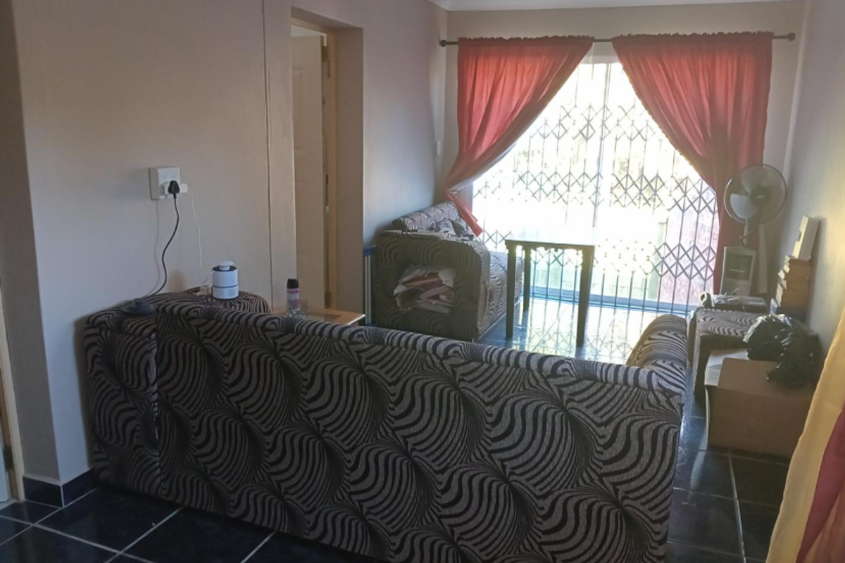 A living room featuring dark floor tiles, a sliding glass door with security gate, and access to an adjacent room.