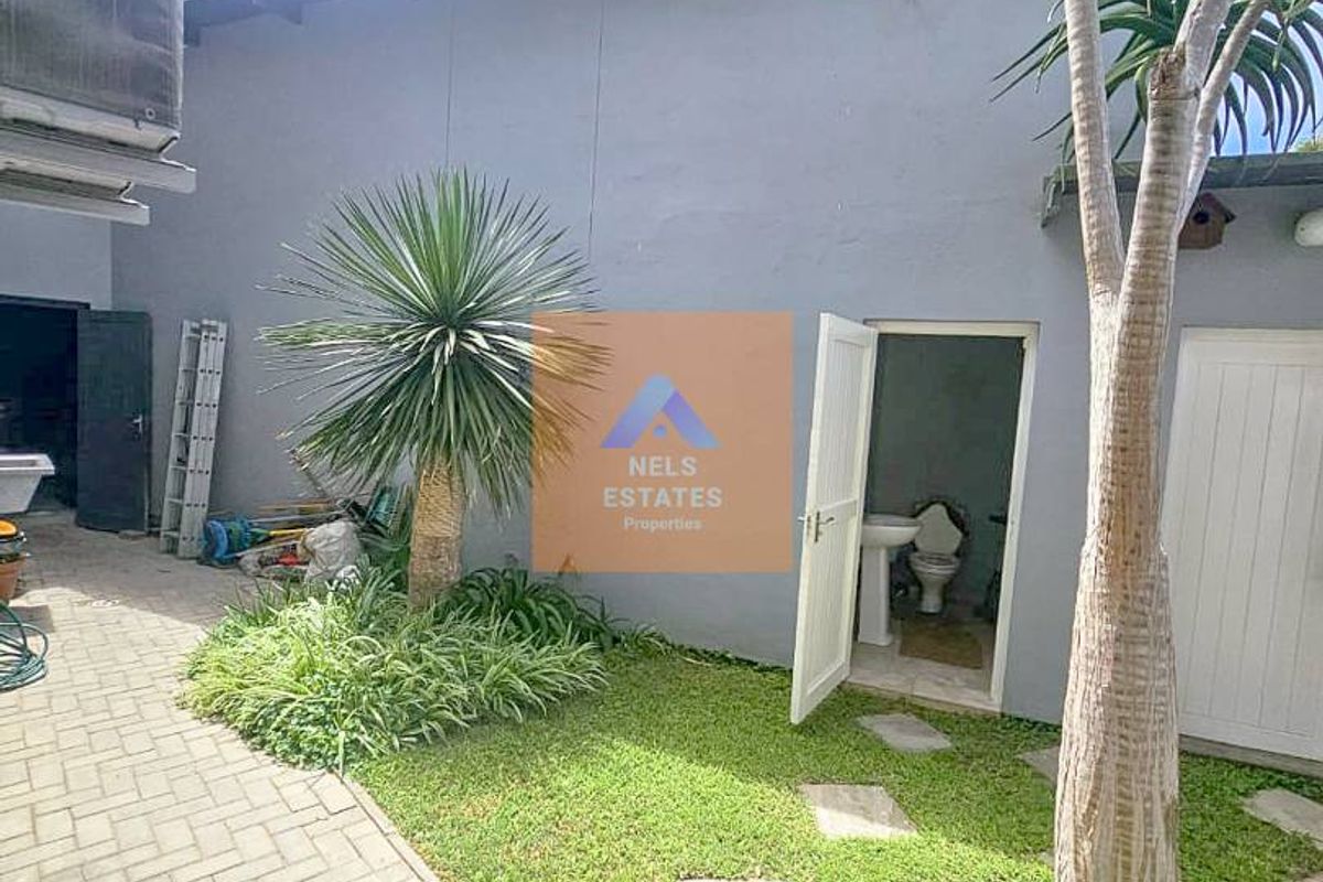 Rear exterior view featuring a paved courtyard, landscaped garden area, and access to an outdoor bathroom facility.