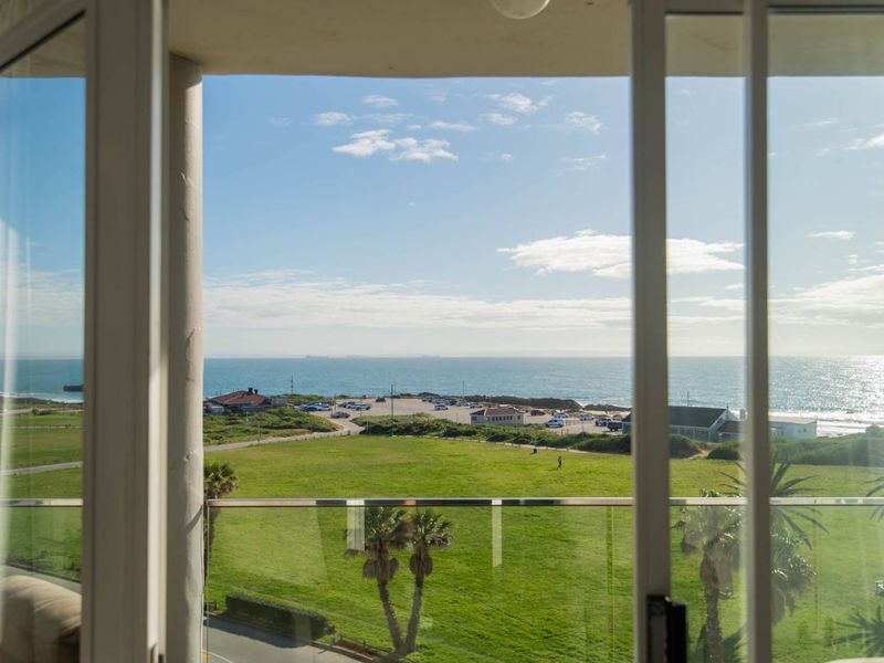 A scenic view of the coastline and green parkland captured through a glass-balustrade balcony and sliding door frame.