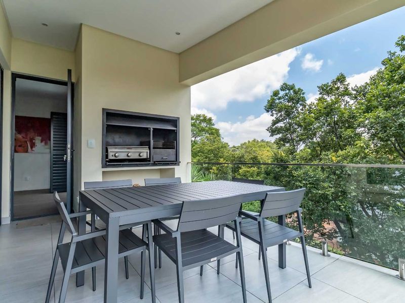Covered balcony featuring a built-in braai, modern glass balustrade, and tiled flooring, offering a pleasant outdoor entertainment space with leafy views.