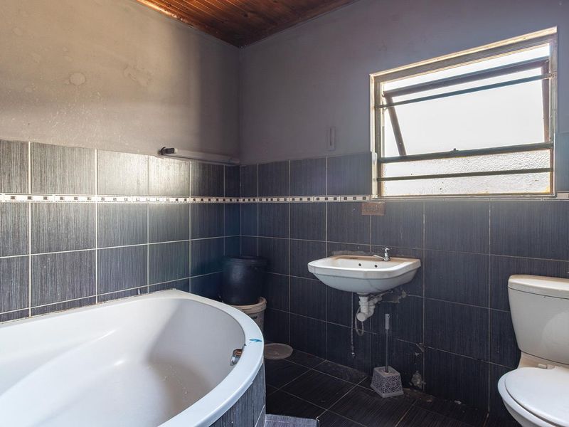 A dated bathroom featuring a bathtub, wall-mounted basin, and toilet, with dark tiled finishes and a timber ceiling, requiring cosmetic updates.