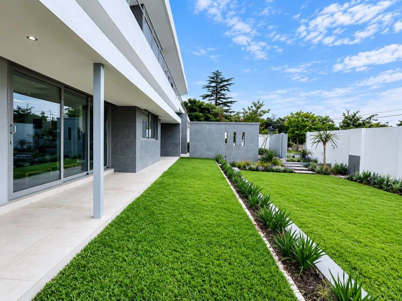 A modern rear exterior featuring a spacious tiled patio, large sliding glass doors, and a meticulously landscaped garden with manicured lawn and retaining walls.