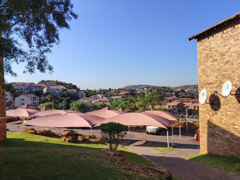 View of the townhouse complex exterior featuring brick facade, paved driveway, and covered parking bays with shade cloth structures.