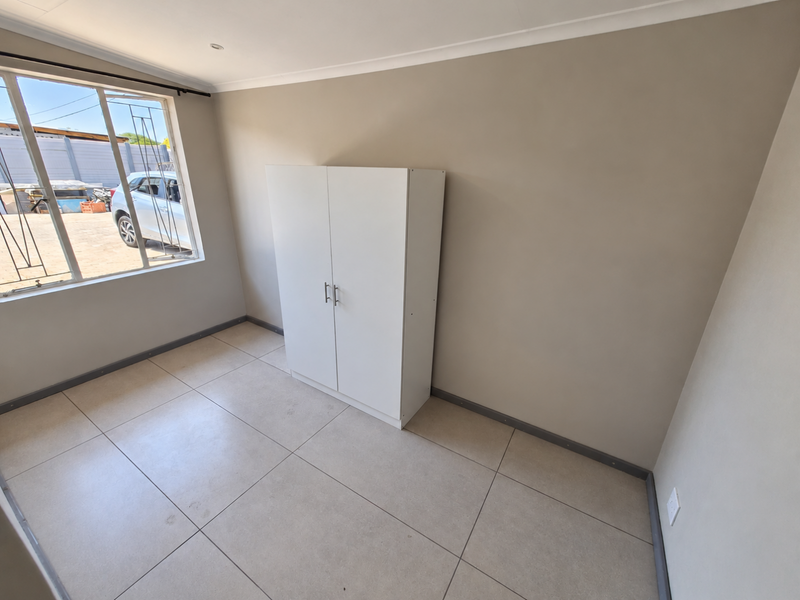 A clean, neutral bedroom featuring large format floor tiles, a built-in cupboard, and security bars on the window.