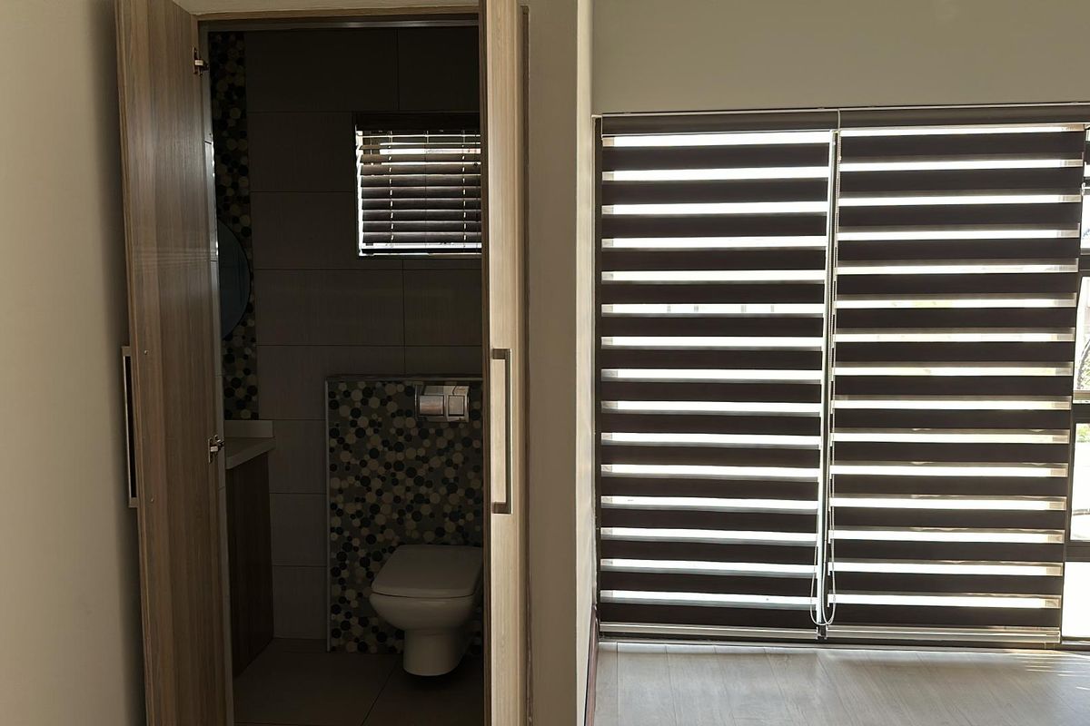 A bright bedroom featuring laminate flooring, modern roller blinds, and an en-suite bathroom with decorative mosaic tiling.