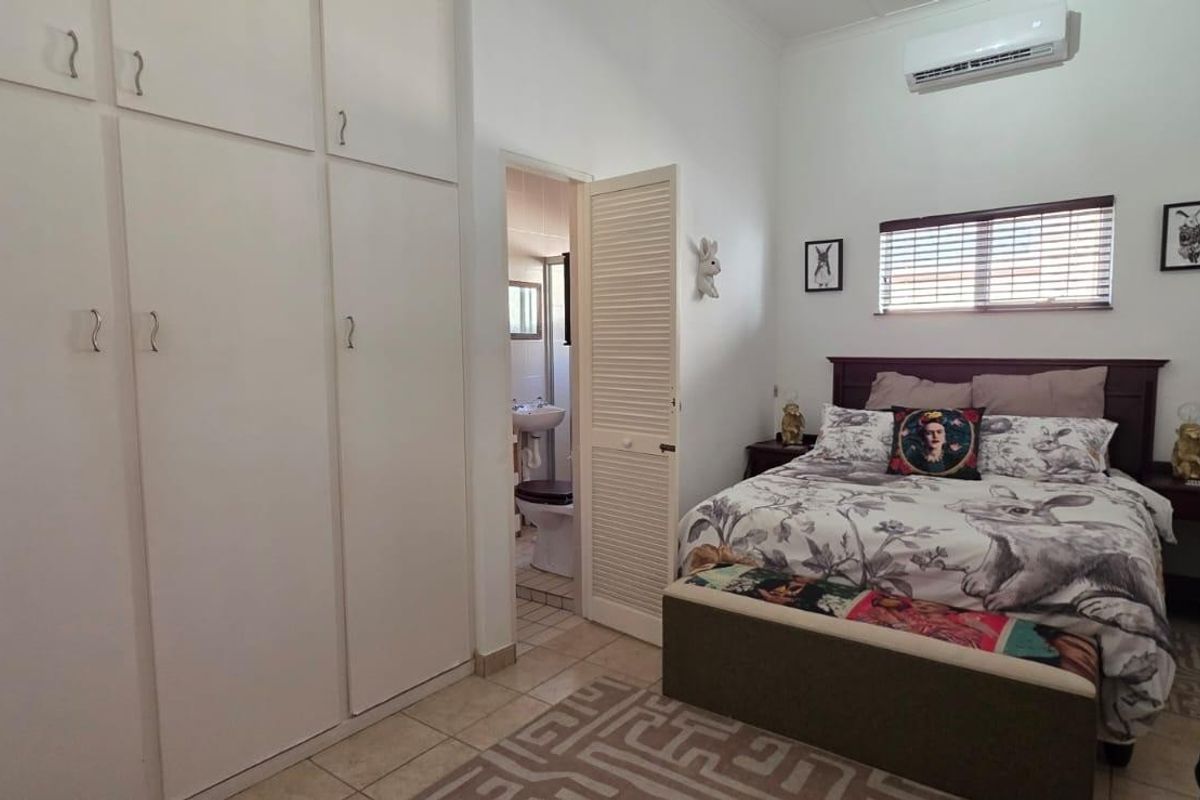 A bedroom featuring built-in cupboards, tiled flooring, and a wall-mounted air conditioning unit, with an adjoining en-suite bathroom visible through the doorway.