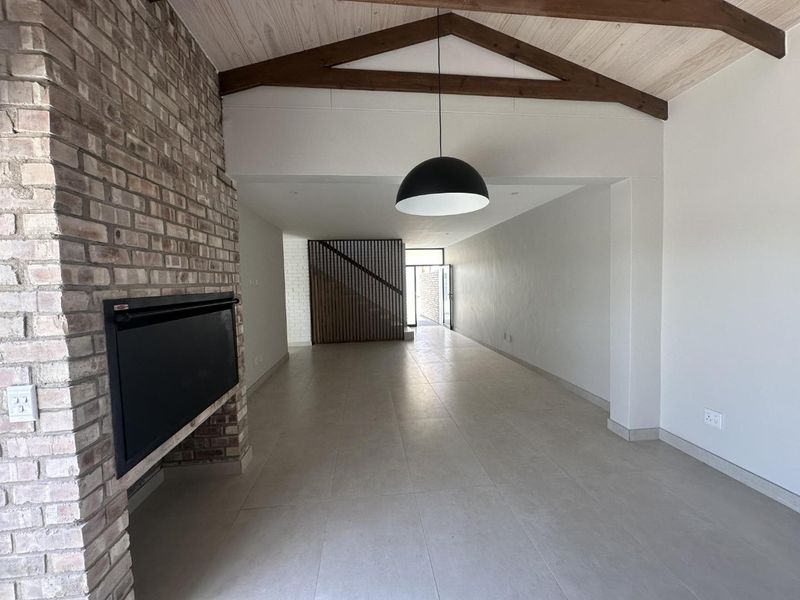 Spacious open-plan living area featuring a striking exposed brick fireplace, vaulted ceilings with timber beams, and modern tiled flooring.