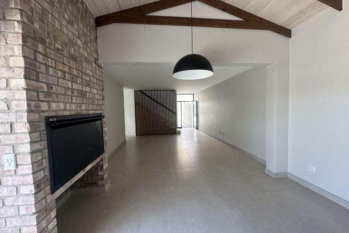 Spacious open-plan living area featuring a striking exposed brick fireplace, vaulted ceilings with timber beams, and modern tiled flooring.