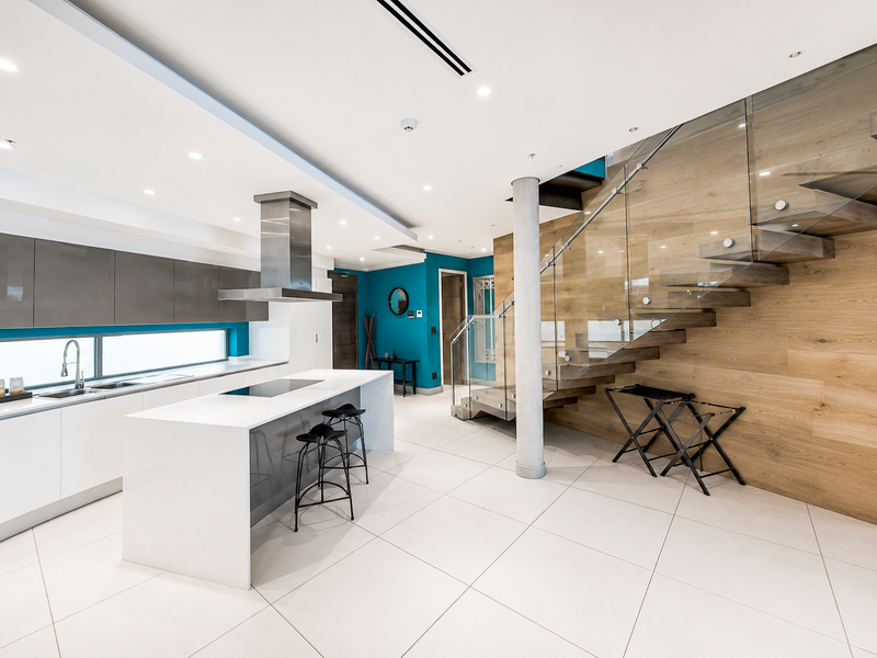 Modern open-plan kitchen featuring a sleek island, high-end cabinetry, and a striking glass-railed staircase with timber wall accents in a contemporary penthouse setting.