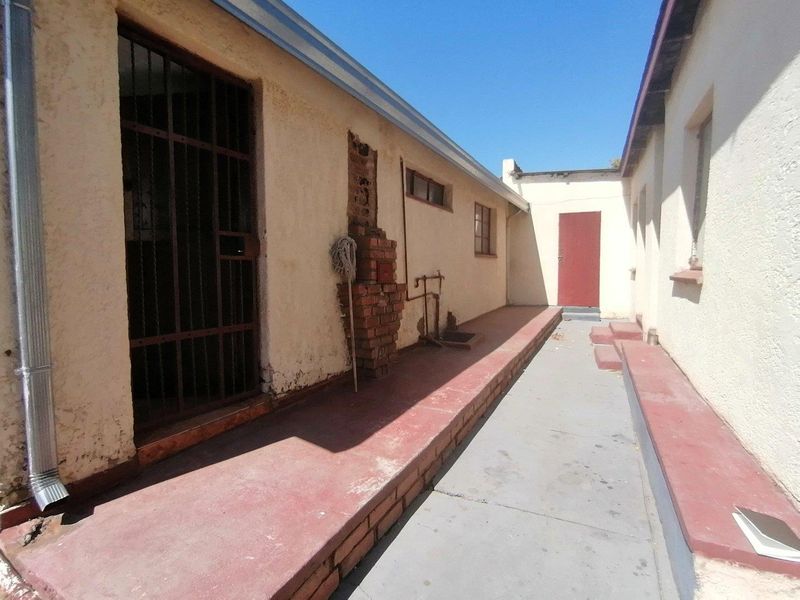 Rear exterior walkway featuring painted concrete surfaces, security gate, and exposed brickwork, showing signs of significant weathering and maintenance requirements.