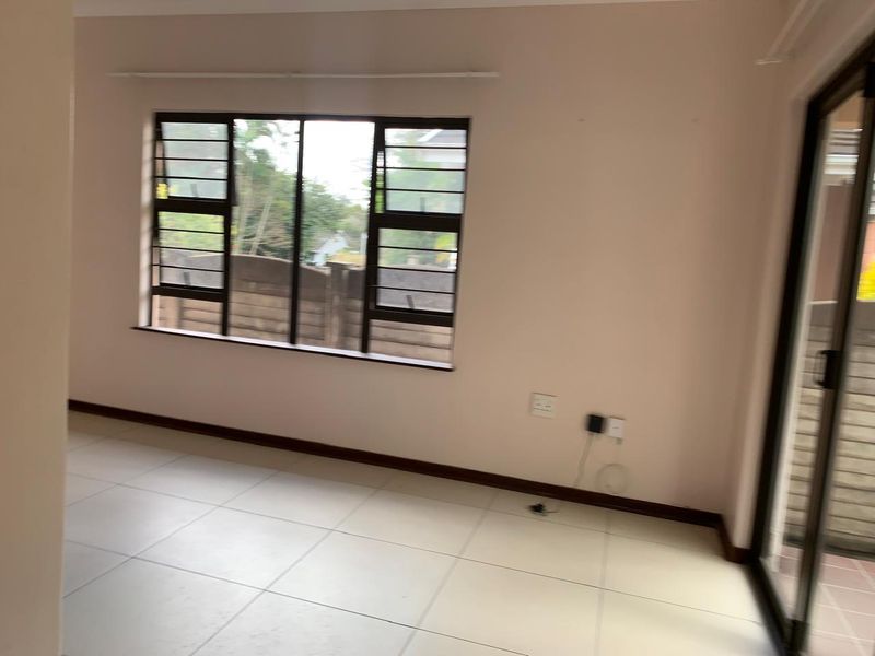 An empty living room featuring neutral tiled flooring, aluminium window frames with security bars, and a sliding glass door leading to the exterior.