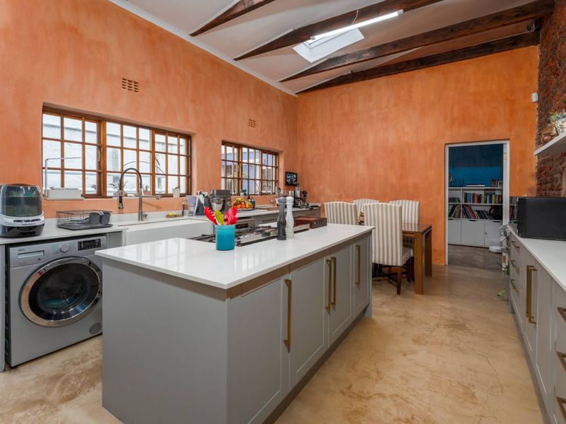 A modern kitchen featuring a central island with quartz countertops, farmhouse sink, gas cooktop, exposed timber beams, and a striking orange feature wall.