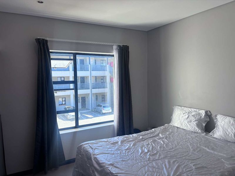 A well-lit bedroom featuring neutral grey walls, a large aluminium-framed window, and recessed ceiling lighting, offering a clean and modern living space.