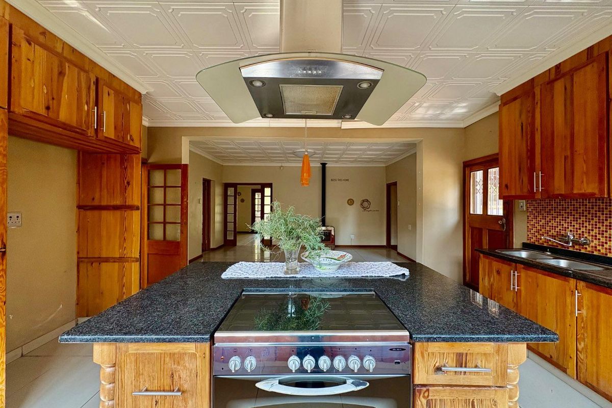 Spacious kitchen featuring a central island with a built-in stove, extractor fan, granite countertops, and warm-toned wooden cabinetry, flowing into an open-plan living area.