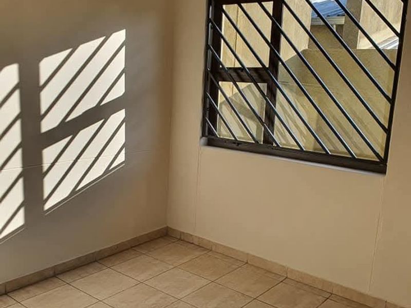 An empty bedroom featuring neutral tiled flooring, a window with security bars, and a curtain rail, offering a clean and versatile living space.