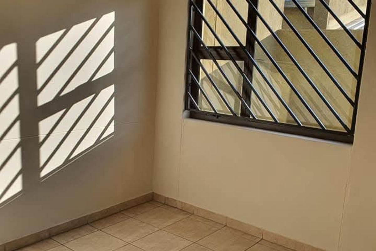 An empty bedroom featuring neutral tiled flooring, a window with security bars, and a curtain rail, offering a clean and versatile living space.