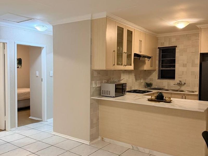 Open-plan kitchen featuring light wood cabinetry, marble-look countertops, and tiled flooring, seamlessly connecting to the adjacent living area and hallway.