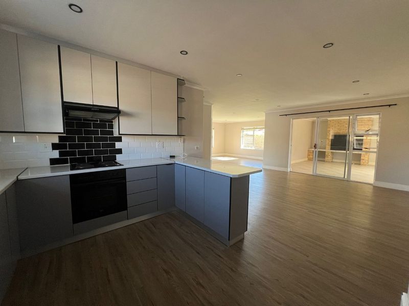 Spacious open-plan kitchen and living area featuring modern two-tone cabinetry, laminate flooring, and seamless flow to an enclosed patio with a brick fireplace.