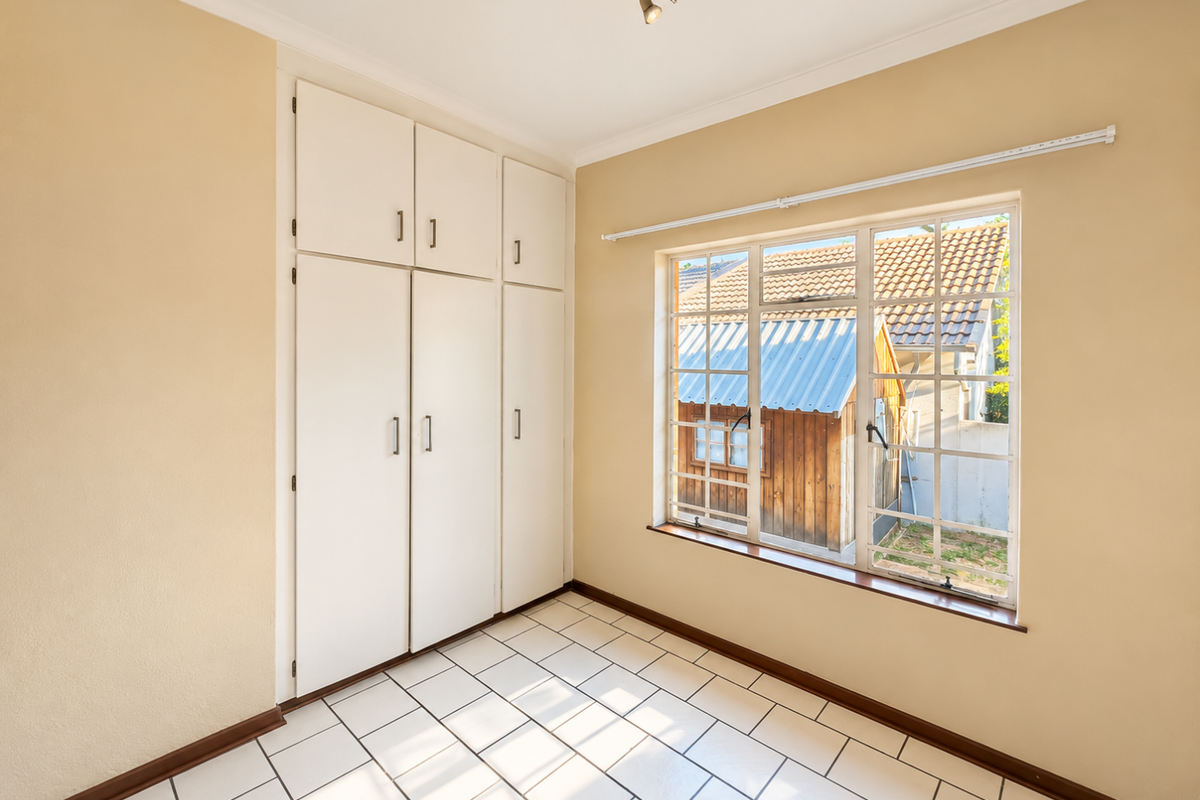 A bright, empty bedroom featuring built-in cupboards, tiled flooring, and a window overlooking the rear garden area.