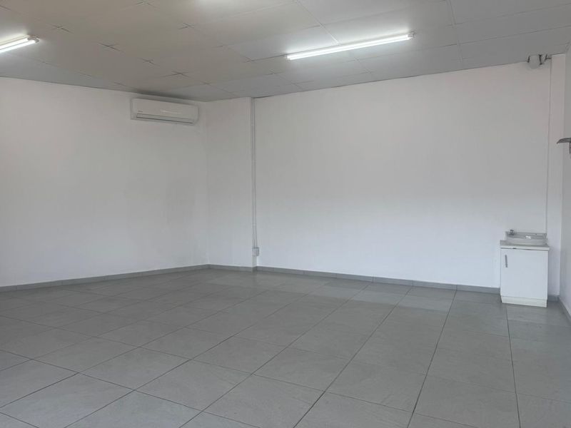 A clean, open-plan retail space featuring tiled flooring, a suspended ceiling, integrated lighting, a wall-mounted air conditioning unit, and a small vanity sink.