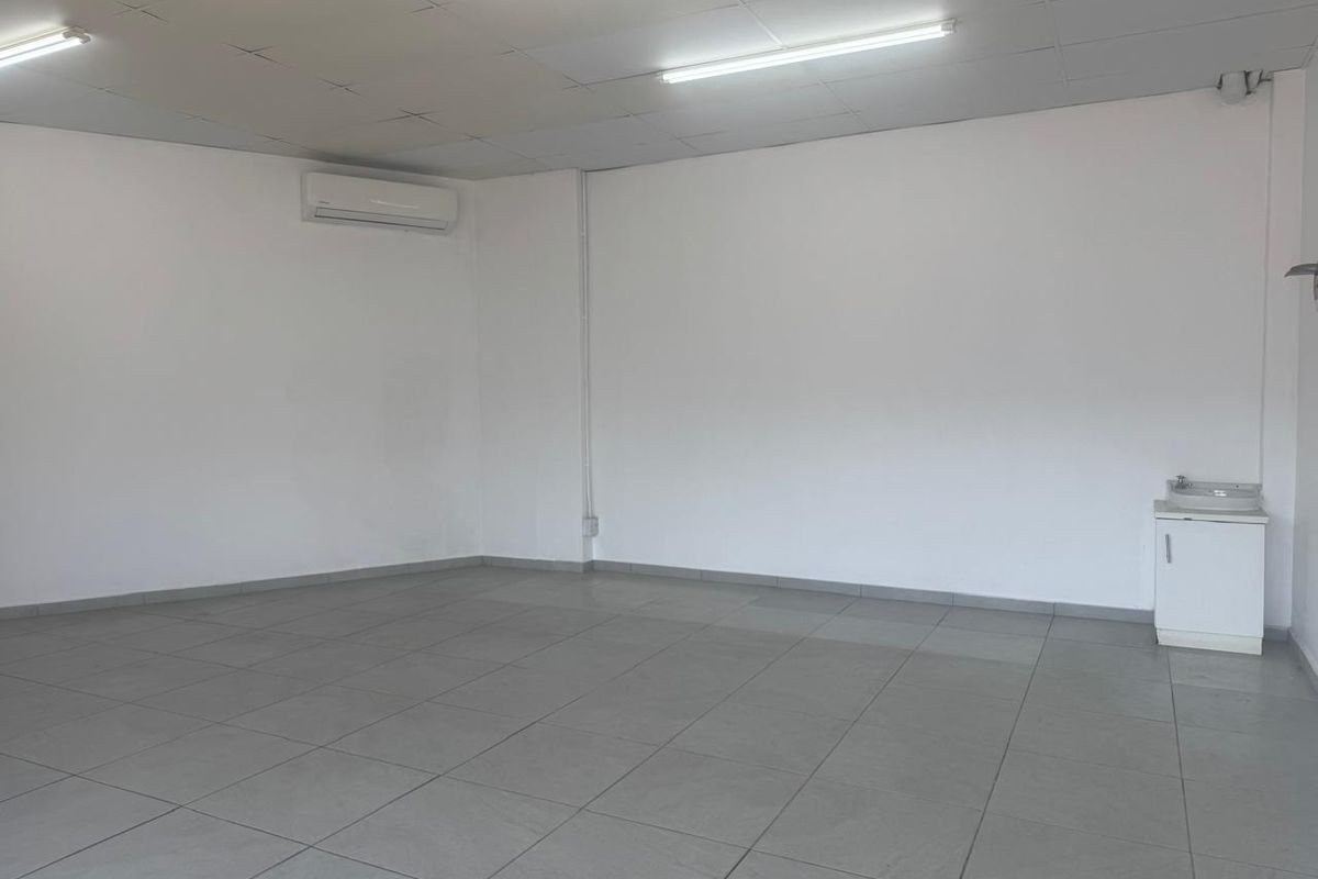 A clean, open-plan retail space featuring tiled flooring, a suspended ceiling, integrated lighting, a wall-mounted air conditioning unit, and a small vanity sink.