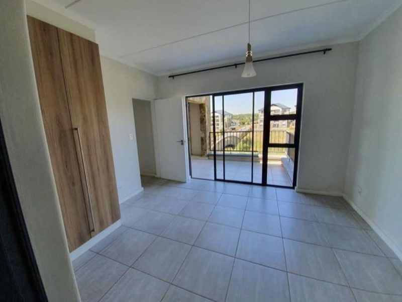 A bright, empty bedroom featuring neutral tiled flooring, a modern built-in wardrobe, and a sliding glass door leading to a private balcony.