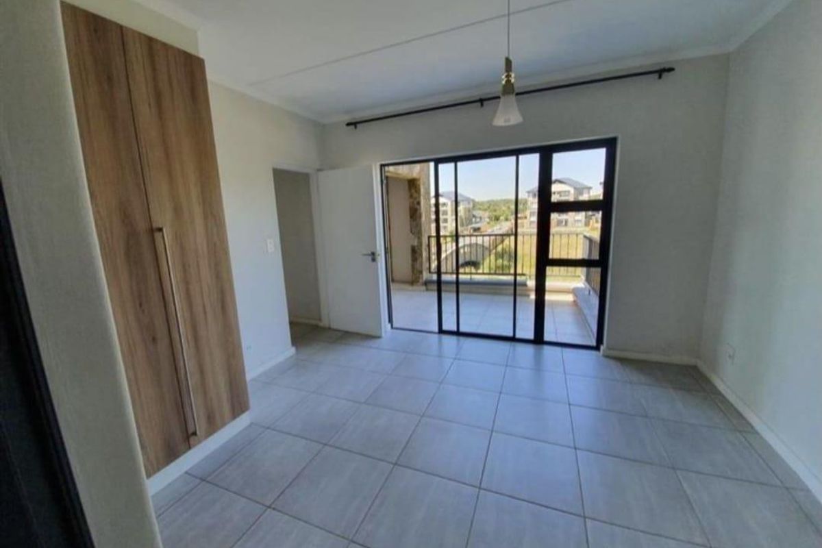 A bright, empty bedroom featuring neutral tiled flooring, a modern built-in wardrobe, and a sliding glass door leading to a private balcony.