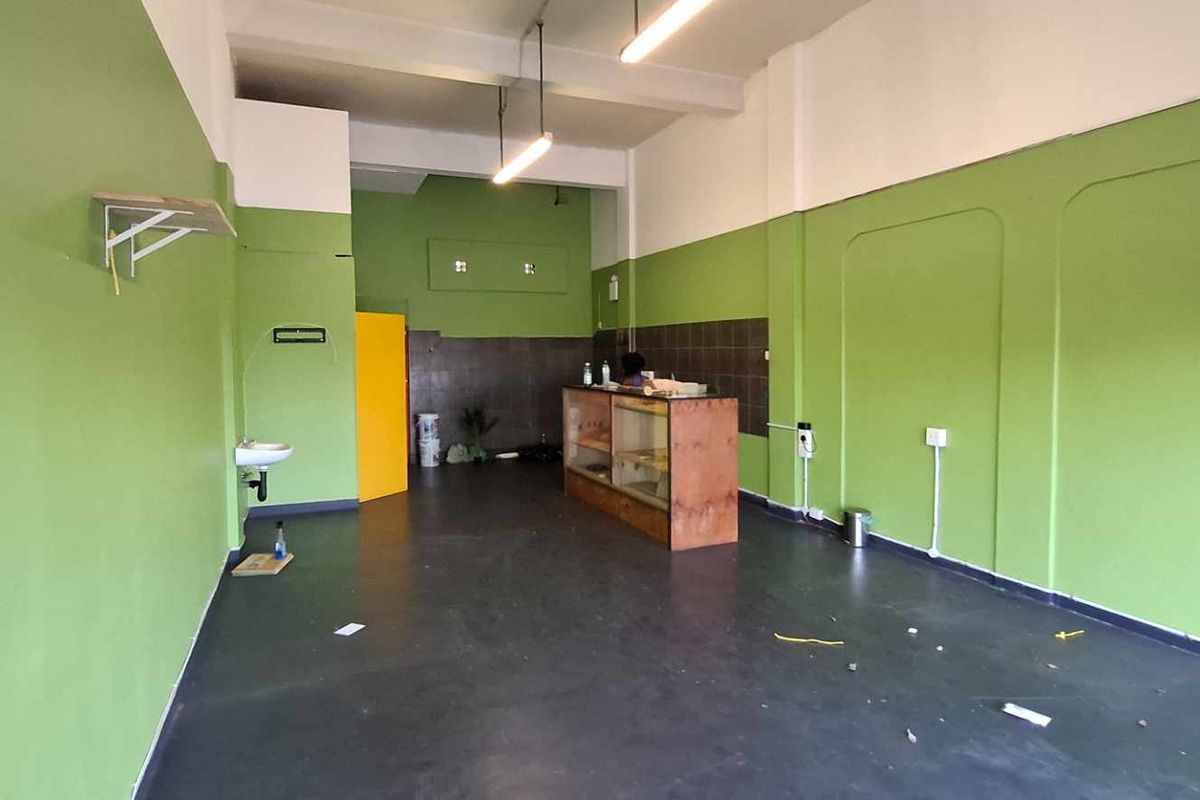 An open-plan commercial space featuring green painted walls, exposed ceiling beams, a small wall-mounted basin, and tiled service area.