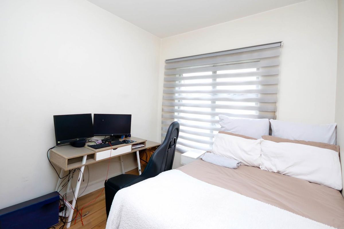 A functional bedroom featuring laminate flooring and modern roller blinds, currently utilized as a combined sleeping and workspace area.