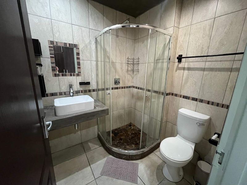 A compact, modern bathroom featuring a corner shower enclosure, vessel sink on a concrete-style vanity shelf, and neutral wall tiling.