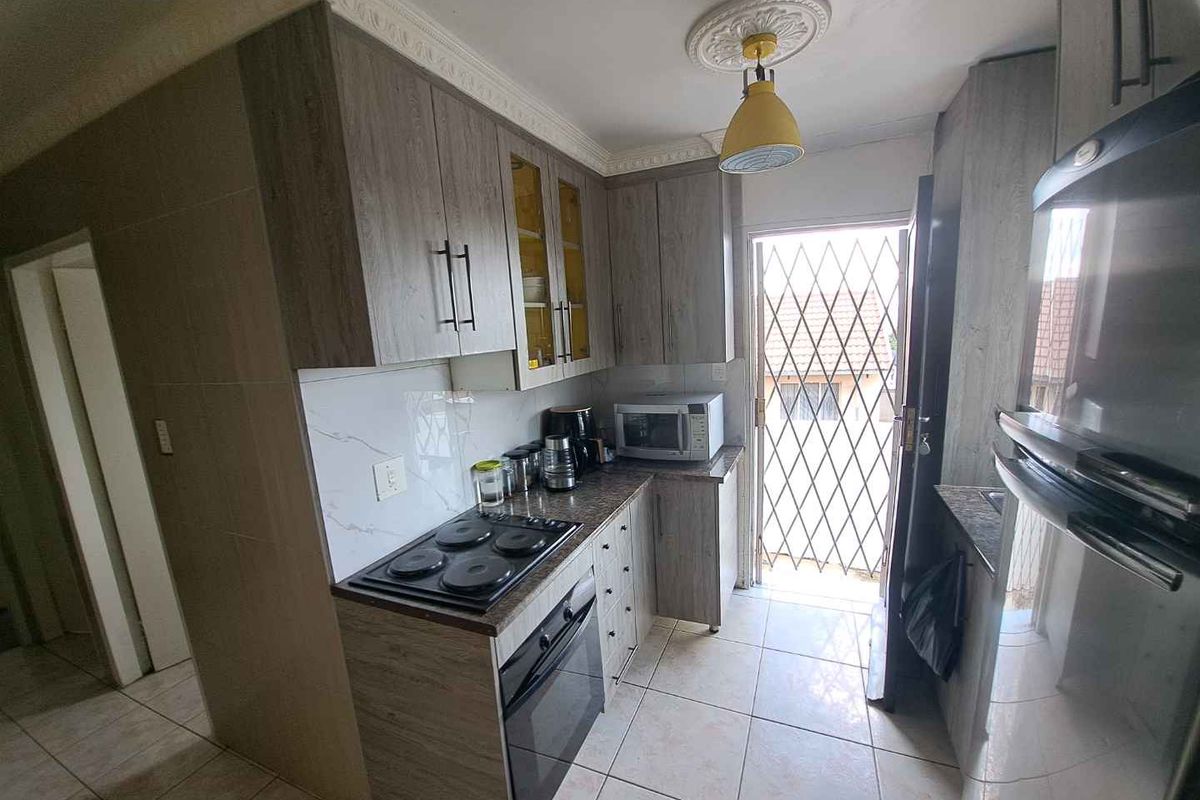 Compact kitchen featuring wood-look cabinetry, built-in oven and hob, tiled flooring, and direct access to the exterior via a security gate.