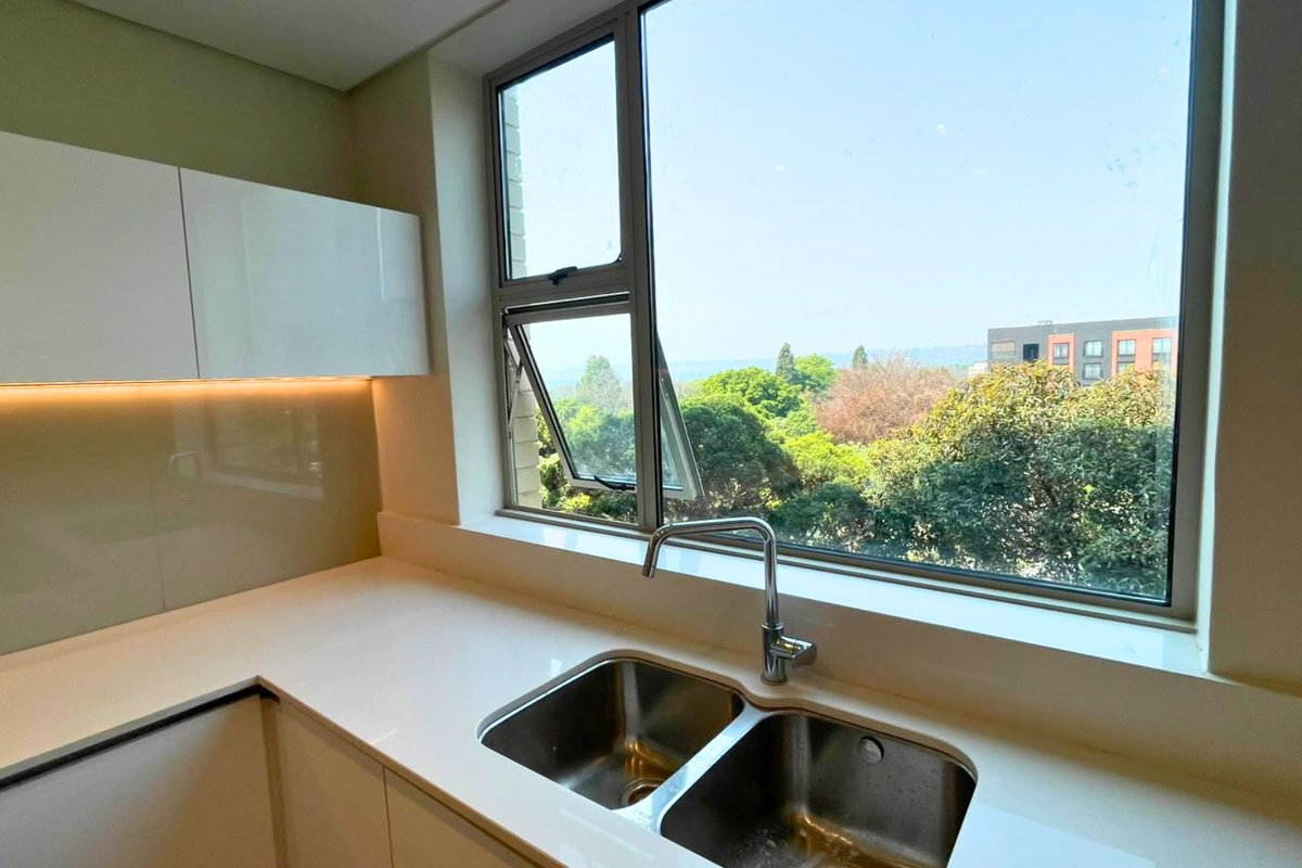 Modern kitchen area featuring sleek white cabinetry, quartz countertops, a double stainless steel sink, and a large window with a pleasant green outlook.
