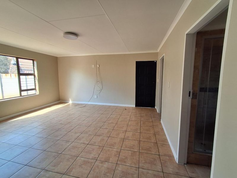 A spacious, empty living area featuring neutral wall tones, tiled flooring, and natural light from a large window, with direct access to an adjacent room.