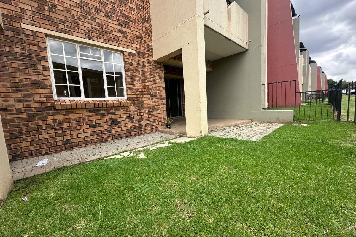 A ground-level exterior view of an apartment unit featuring a private paved patio area, brick facade, and access to a shared garden space.