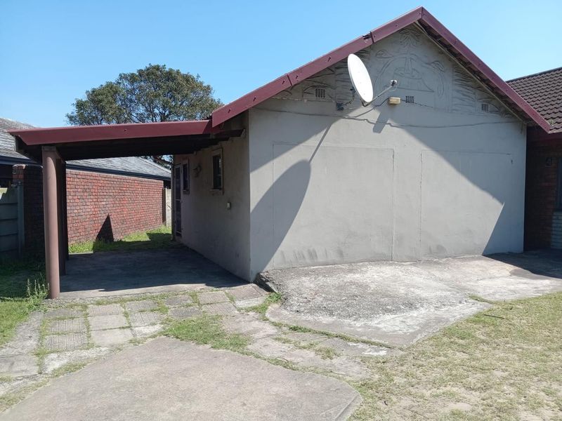 Rear view of the property featuring a carport, concrete driveway, and brick boundary wall, showing signs of weathering and wear.