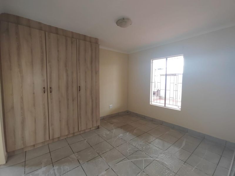 A clean, empty bedroom featuring built-in wooden cupboards, tiled flooring, and a window fitted with security bars.