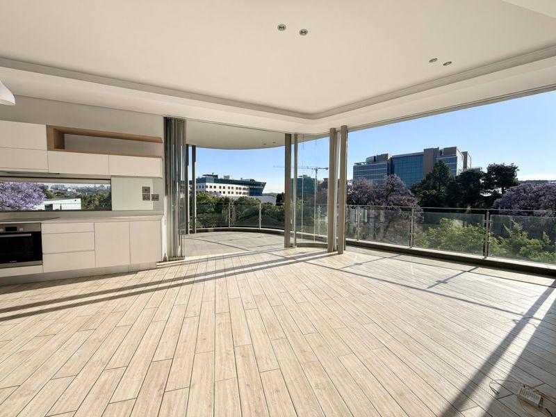 Spacious open-plan living area featuring modern kitchen cabinetry, wood-look tile flooring, and expansive glass sliding doors opening onto a private balcony with city views.