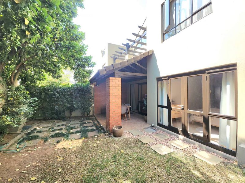 A rear exterior view featuring a covered patio with a brick pillar, timber pergola, and direct access to a private garden with paved pathways.