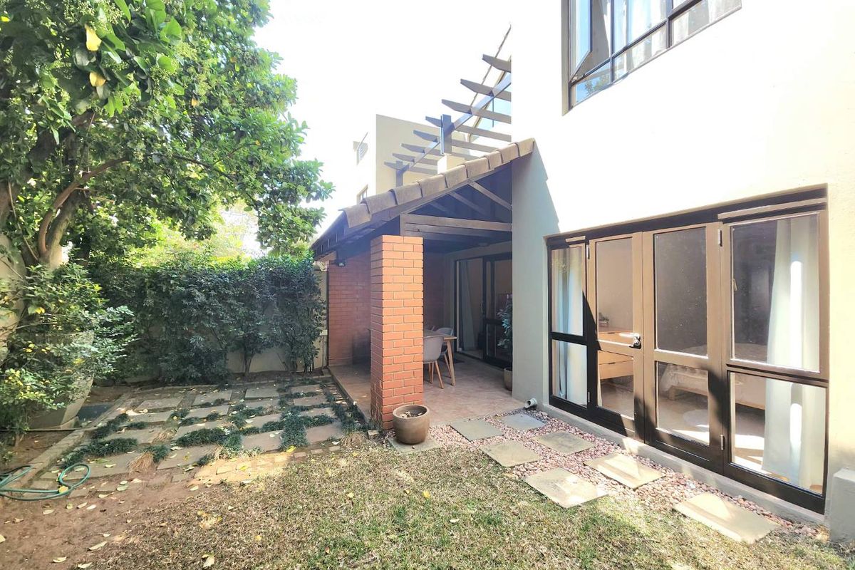 A rear exterior view featuring a covered patio with a brick pillar, timber pergola, and direct access to a private garden with paved pathways.