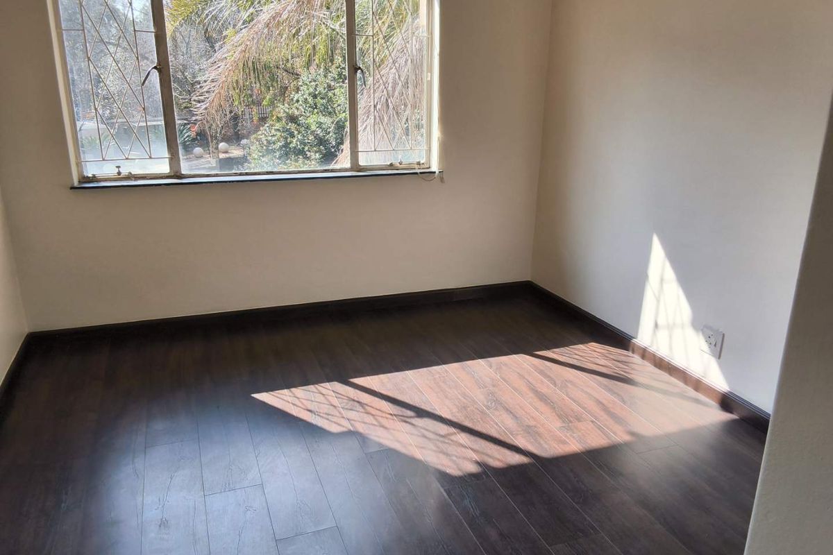 A bright, empty bedroom featuring dark wood-look laminate flooring and a large window with security bars overlooking the garden.