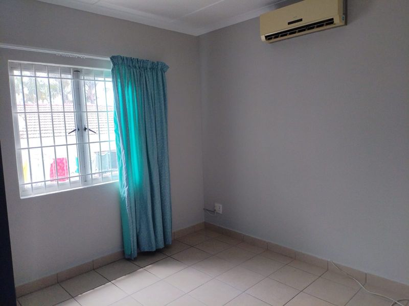 A clean, empty bedroom featuring tiled flooring, a wall-mounted split-system air conditioner, and a window fitted with security bars.